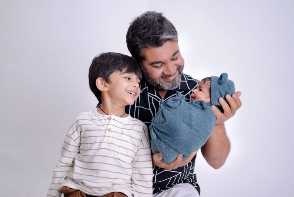 A dad and older sibling smiling at a newborn baby wrapped in a blue swaddle.