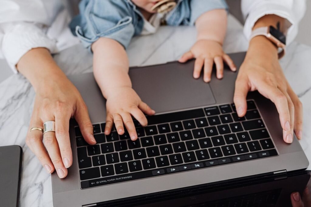 A baby's hands resting on a laptop keyboard next to a parent's hands, representing balancing work and baby.