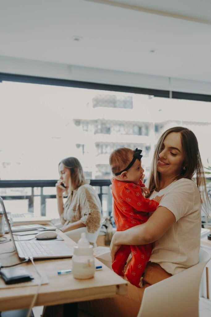 A mother holding her baby in an office setting while a colleague works nearby.